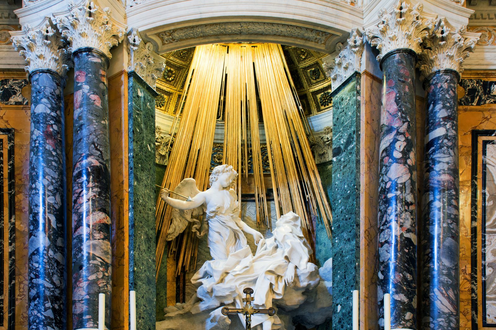 Marble sculpture by Bernini of Saint Teresa of Ávila in a swoon of mystical ecstasy, with an angel above her holding a golden arrow, set within a niche of gilded light.